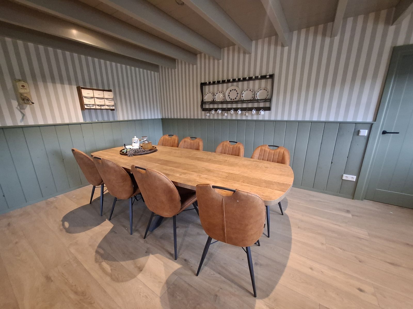 De eetkamer met een lange houten tafel voor acht personen, gestreept behang en saliegroene lambrisering